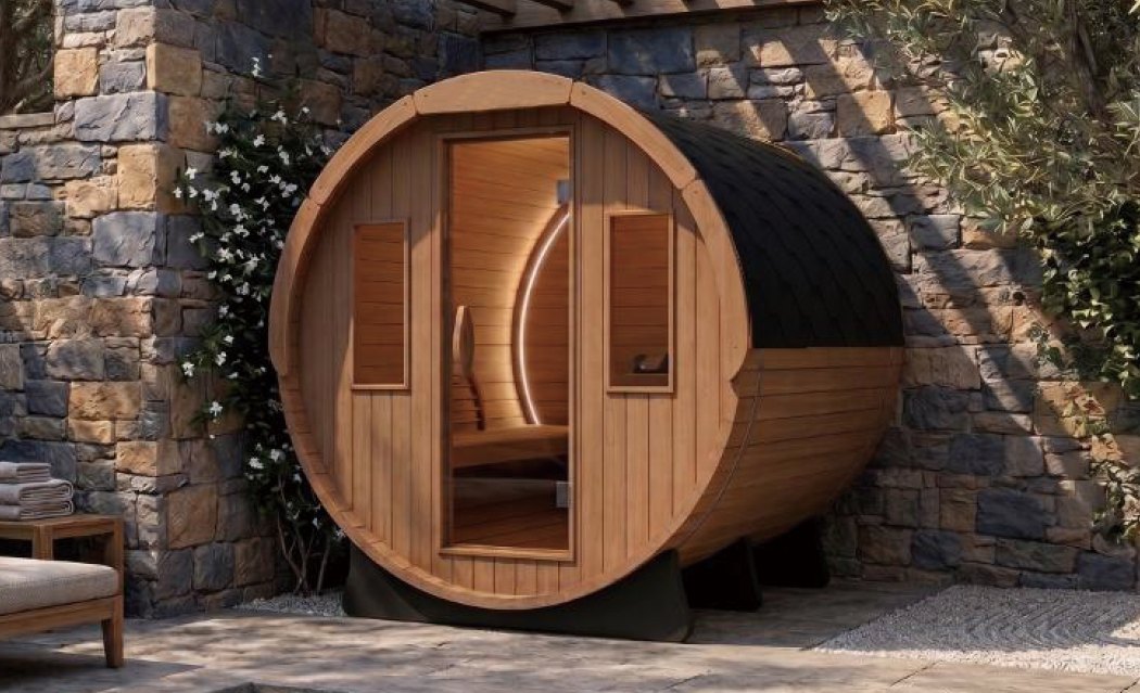 Sauna barril EREZ Brasa junto a parede de pedra, com jasmim florido e telhado escuro.